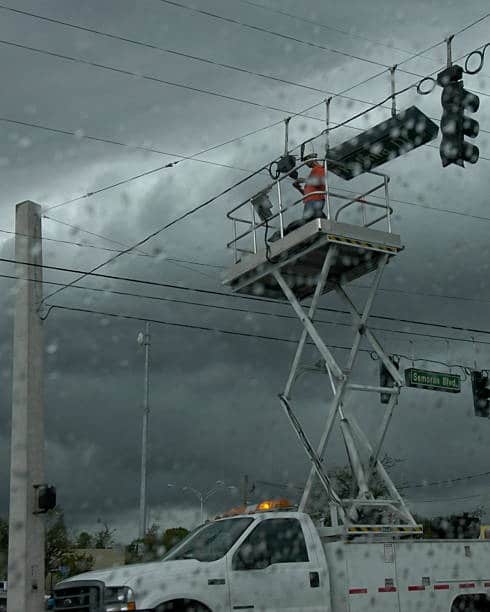 Lineman in a lift repairing traffic lights during a storm, viewed through a rain-covered window.